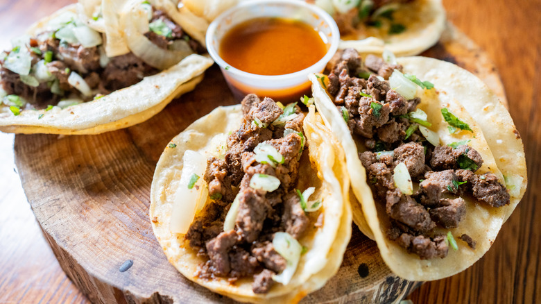 Four Carne Asada tacos on a wooden board with an orange dipping sauce in the center