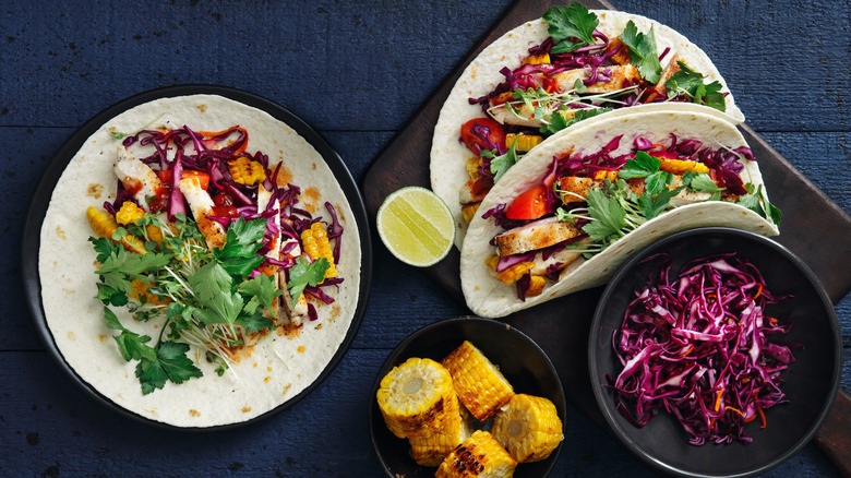 Grilled chicken tacos with corn, red cabbage and cilantro on a wooden board on top of a dark bl;ue background. A bowl of corn and cabbage sits to the side