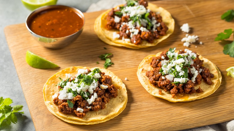 Three chorizo tacos topped with cheese and cilantro on a wooden chopping board decorated with limes and cilantro