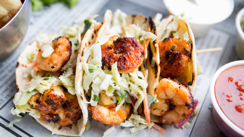 Shrimp tacos topped with slaw on a newspaper, surrounded by dipping post of red, white and green sauce