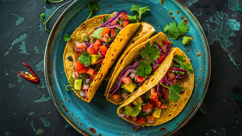 Veggie tacos with tomato, onion, avocado and cilantro on a turquoise place that sits on a turquoise blue background