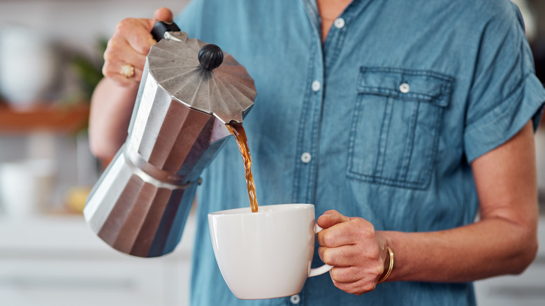 Person pouring coffee out of a moka pot into a white ceramic coffee mug