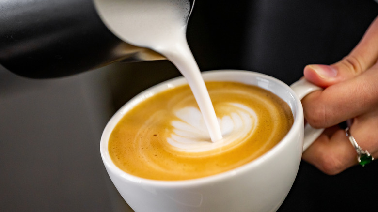 Steamed milk being poured from a metal container into a white ceramic cup