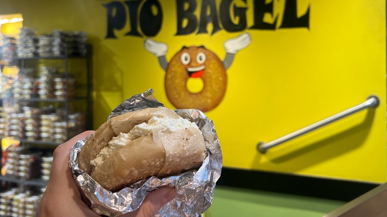 A Pio Bagel gluten-free cream cheese bagel is held in front of the store's logo.