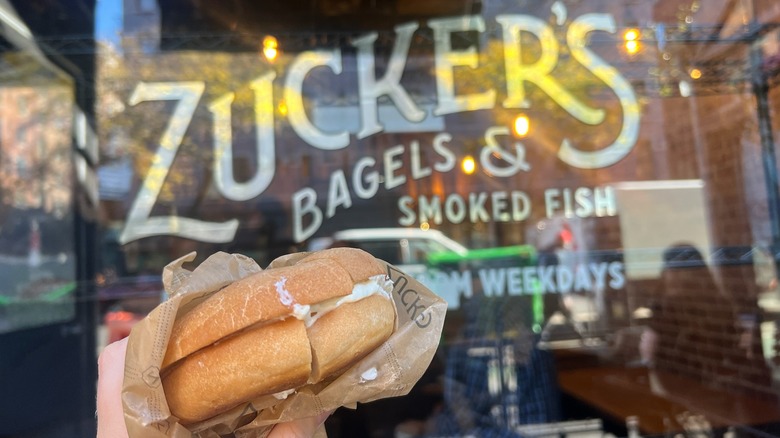 A Zucker's cream cheese gluten-free bagel is held in front of the store.