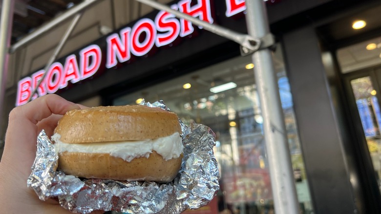 A Broad Nosh gluten-free cream cheese bagel is held in front of the store.