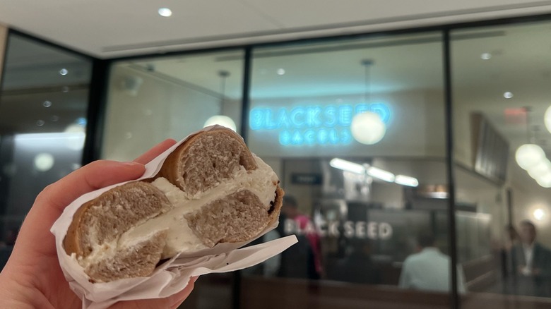 A Black Seed Bagels gluten-free cream cheese bagel is held in front of the store.