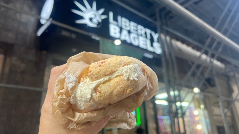 A Liberty Bagels gluten-free cream cheese bagel is held in front of the store.