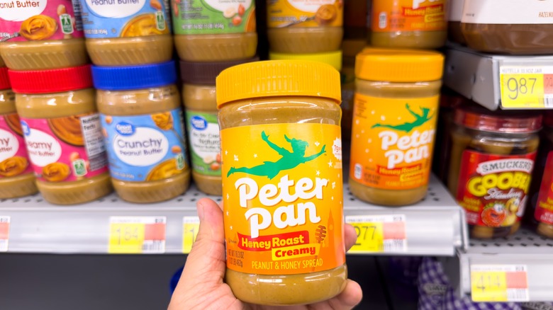 hand holding jar of peter pan peanut butter with shelf of peanut butter jars in background