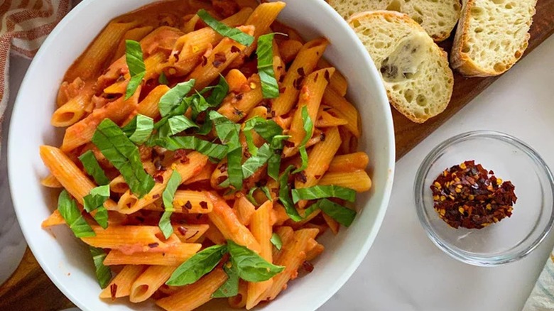 Top-down view of a bowl of penne alla vodka next to bread and pepper flakes