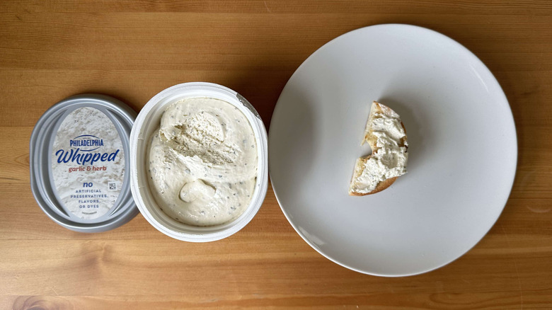 Whipped garlic herb cream cheese is shown on a bagel and in the container.