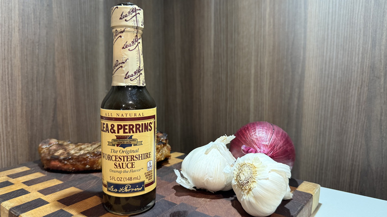 A bottle of Worcestershire sauce with garlic bulbs, red onion, and grilled steak