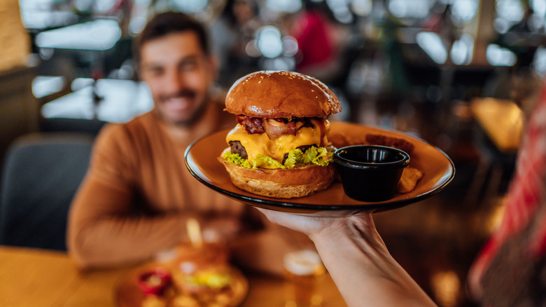 Waiter carrying a brown plate with a burger and a black pot of sauce to a customer in a busy restaurant