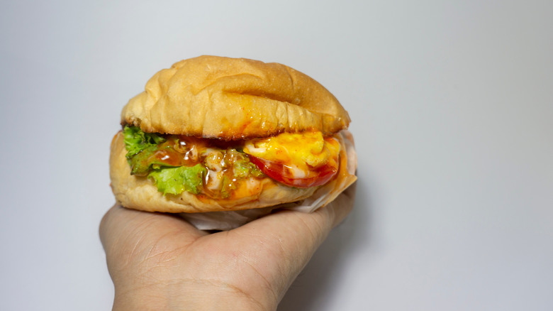 Cheap egg burger with lettuce, tomatoes and sauce in the palm of a persons hand against a grey wall