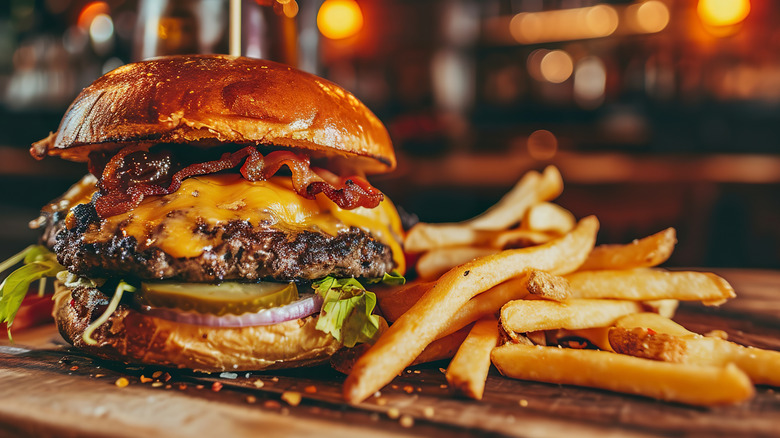 Burger filled a burger patty, bacon, cheese, onion, pickle, lettuce on a wooden board next to a stack of fries