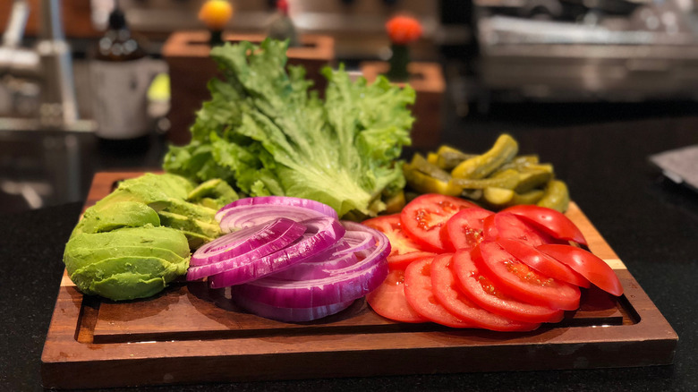 Wooden tray with lettuce, tomato, onion, avocado ad pickles against a background of a kitchen