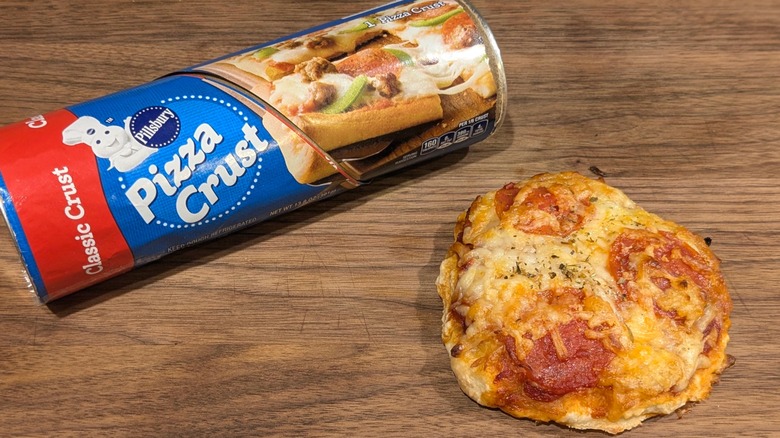 Tube of pizza dough and tiny, thick pizza with pepperoni on wood board