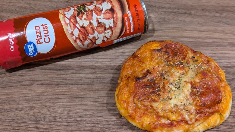 Red tube of pizza dough next to ball-shaped tiny pizza with cheese, herbs, and pepperoni on wood board