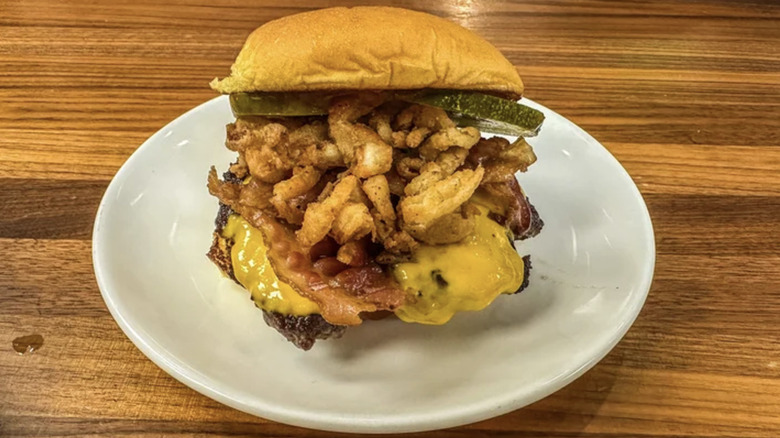 Shake Shack burger coverd in cheese, bacon, pickles, friend onions, on a white plate on a wooden table.