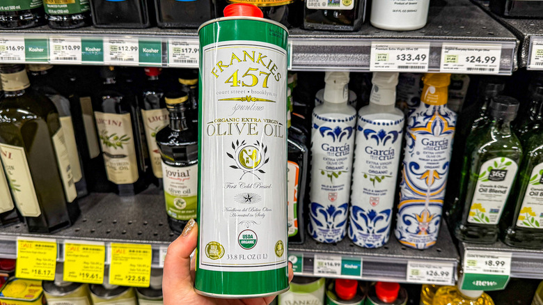 Hand holding bottle of Frankies 457's organic extra virgin olive oil in front of shelves of olive oil