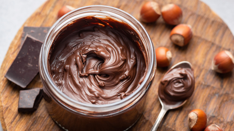 Jar of hazelnut spread with whole hazelnuts and chocolate pieces on the side