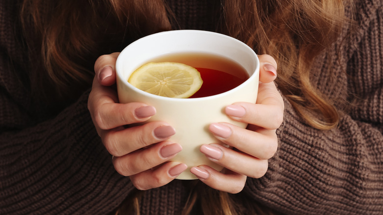 Woman in brown sweater holding white mug of tea with lemon slice