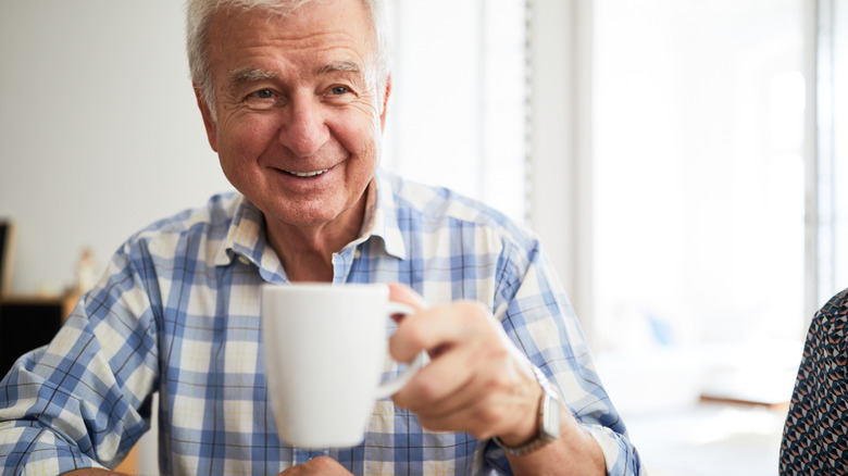 Older man smiles while holding up a white coffee cup