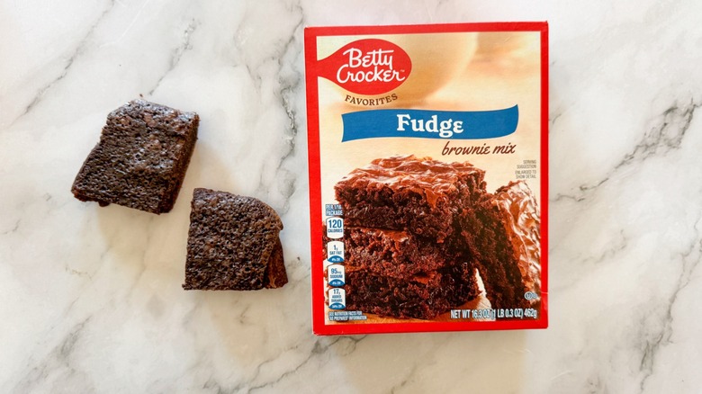 Baked fudge brownie next to boxed brownie mix on marble surface