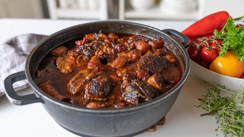 rich braised beef shanks in a cast iron dutch oven, shown on a white countertop next to herbs and a bowl of colorful vegetables