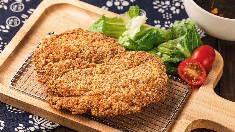beef cutlet (cihcken-fried steak) shown on a cutting board with lettuce and cherry tomato