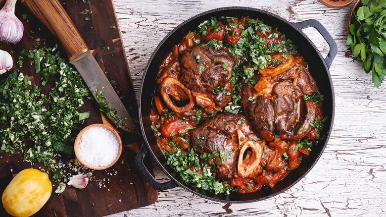 dish of braised osso buco in cast iron pot on rustic white wooden table, next to a cutting board containing fresh-made gremolata of garlic, parsley and lemon