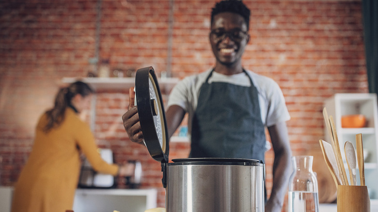 A man opening the lid on a pressure cooker with brick wall behind him