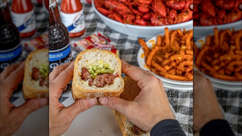 Man's hands holding alligator sausage po'boy with boiled crawfish and french fries in background