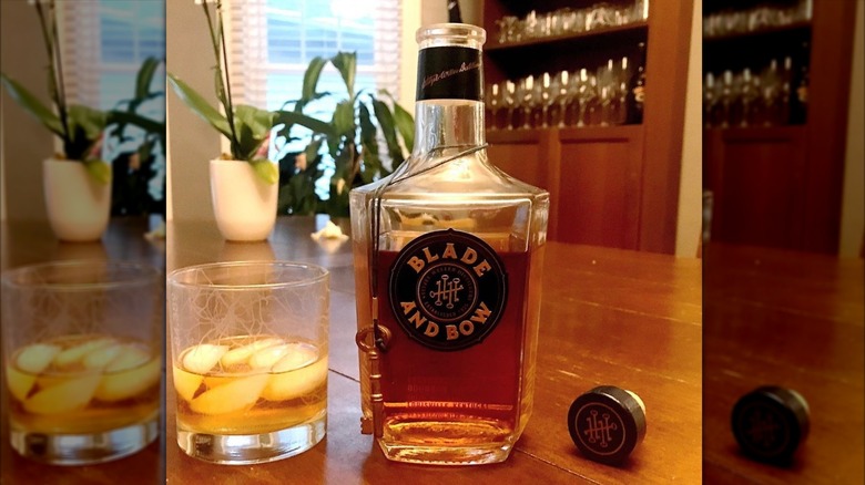 Bottle of Blade and Bow bourbon with a glass filled with bourbon and ice on the side on a wooden table with plants in the back
