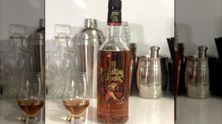 Bottle the Fighting Cock bourbon next to a glass filled with bourbon and cocktail accessories in the back