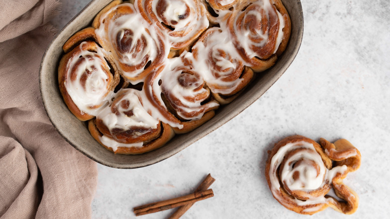 Top view of a tray of cinnamon rolls covered with cream cheese glaze sitting on a marble counter next to a cinnamon roll and cinnamon sticks