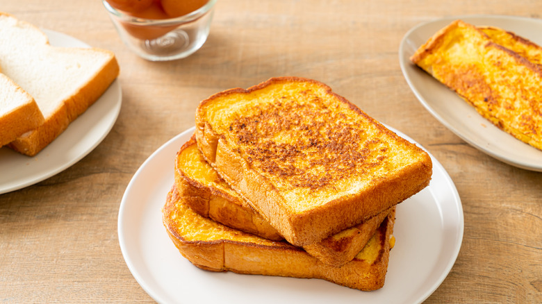 French toast stacked on a white plate on a wooden table with two plates and eggs in the back