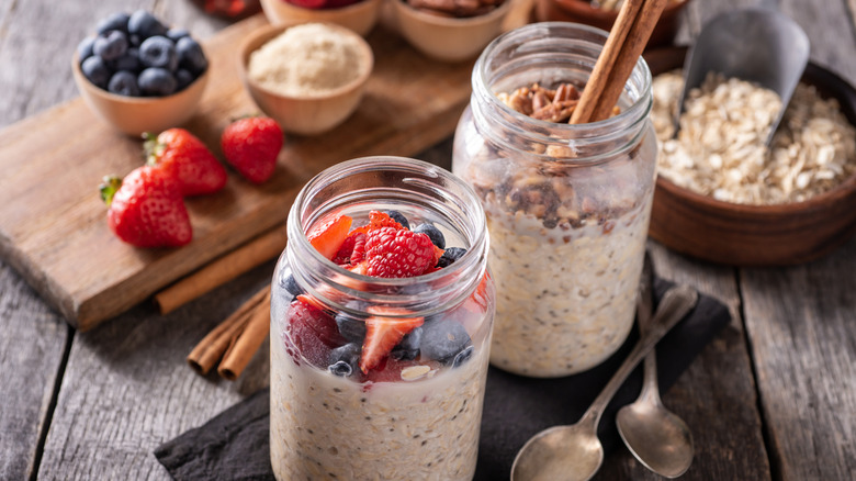 Two jars of overnight oats topped with fresh berries and nuts standing on a wooden table with a cutting board, fresh fruit, and oats in the back