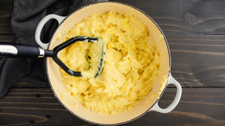 Mashed potatoes being mashed by a potato masher in a white cast iron pot