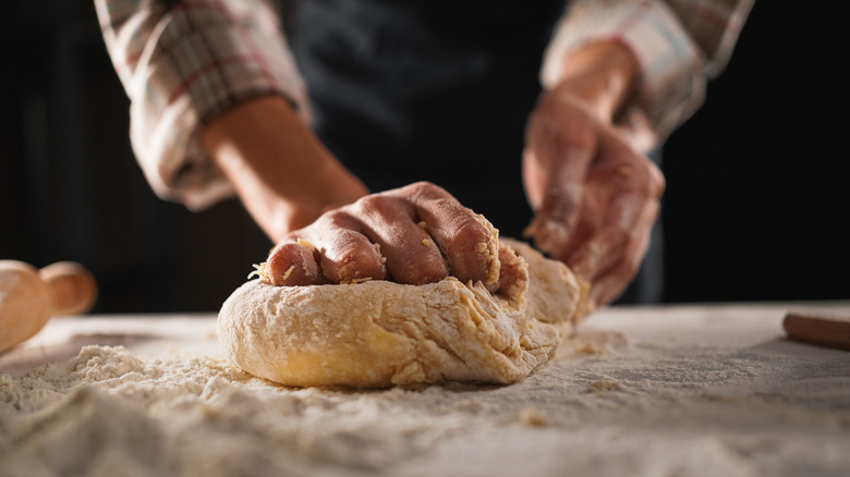 A person kneading bread dough on a flour-coated counter with a rolling pin in the back