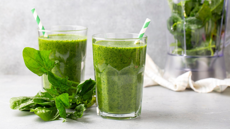 Two tall glasses filled with green smoothies and garnished with paper straws set on the pale background surrounded by fresh spinach