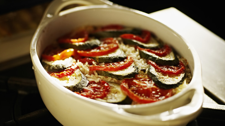 Vegetable casserole assembled in a white casserole dish, topped with tomatoes, zucchinis, and shredded cheese