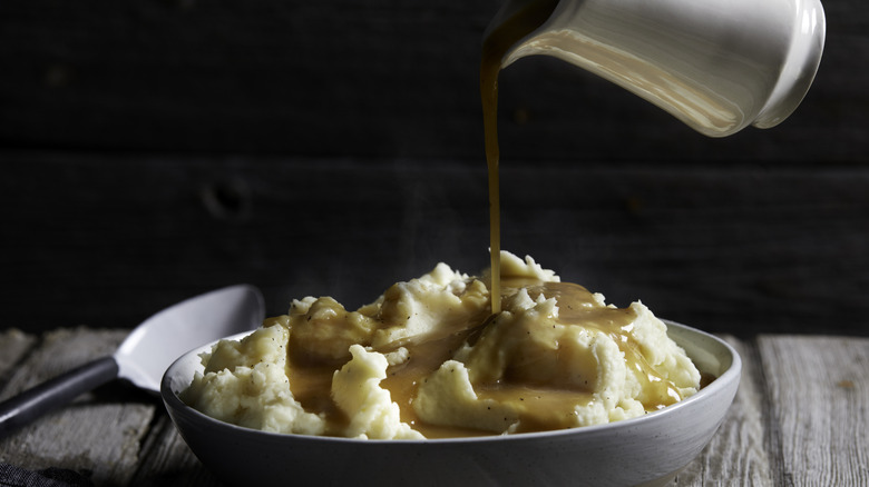 Pouring classic gravy from a white pitcher directly on top of a bowl of creamy mashed potatoes stacked in a white bowl