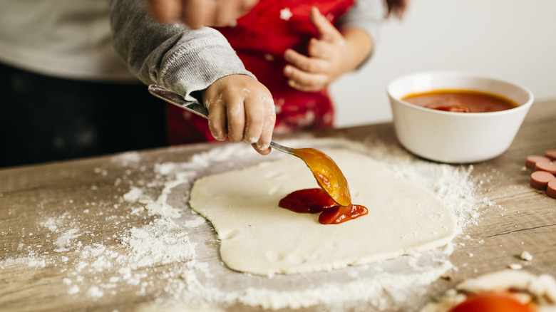 Parent holding a that is spreading a thick tomato sauce on top of fresh pizza dough that's been rolled onto a wooden counter