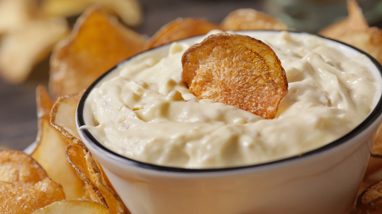 A close up of a French onion dip inside a white bowl with a potato chip as a garnish, surrounded by potato chips with chips