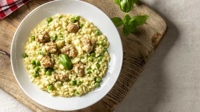 A white bowl sitting on a wooden board filled with a creamy risotto with sausage meat, peas, and fresh basil