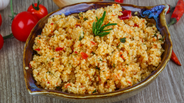 Brown, leaf-shaped ceramic bowl filled with couscous made with tomatoes and herbs, placed on a wood-like surface with fresh tomatoes and dry chilis on the side