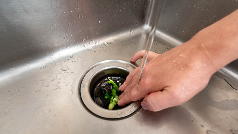 hand pushing food down a garbage disposal in a stainless steel sink with water running