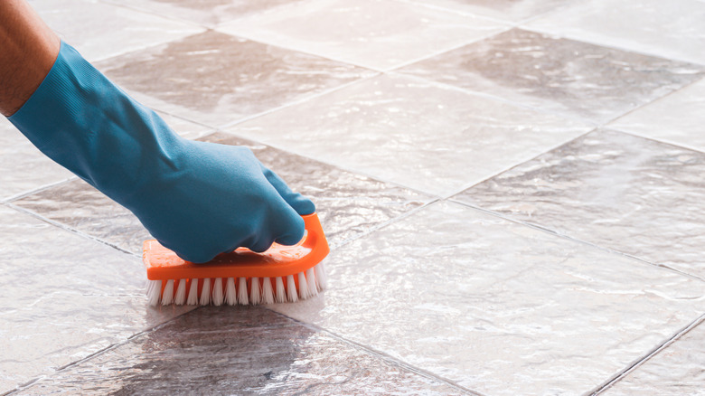 hand with a blue rubber glove scrubbing grout off a beige tile floor