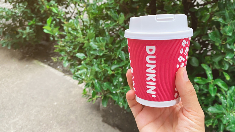 Hand holding a small hot coffee from Dunkin' in front of outdoor foliage